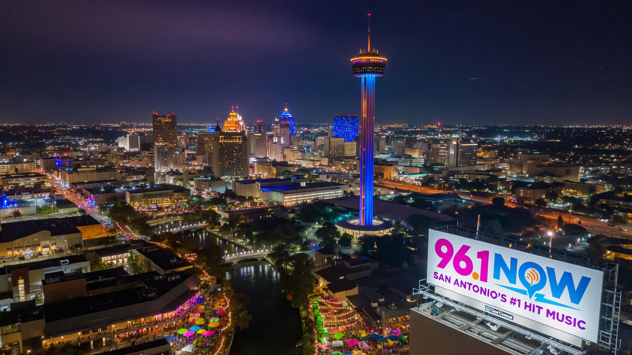 San Antonio Texas city skyline at night with the Tower of the Americas and River Walk lights, a 96.1 NOW billboard in the foreground