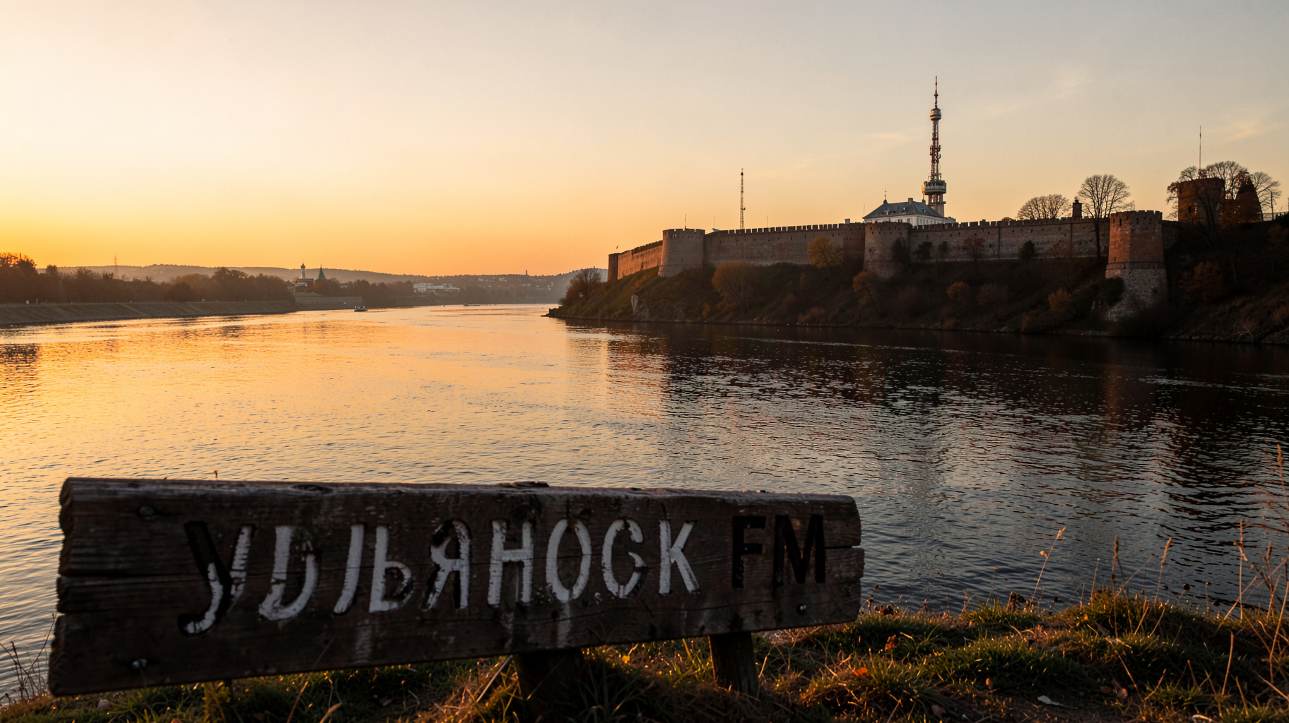 A golden hour view over the Volga river in Ulyanovsk with a fortress silhouette and a wooden sign reading Ульяновск FM
