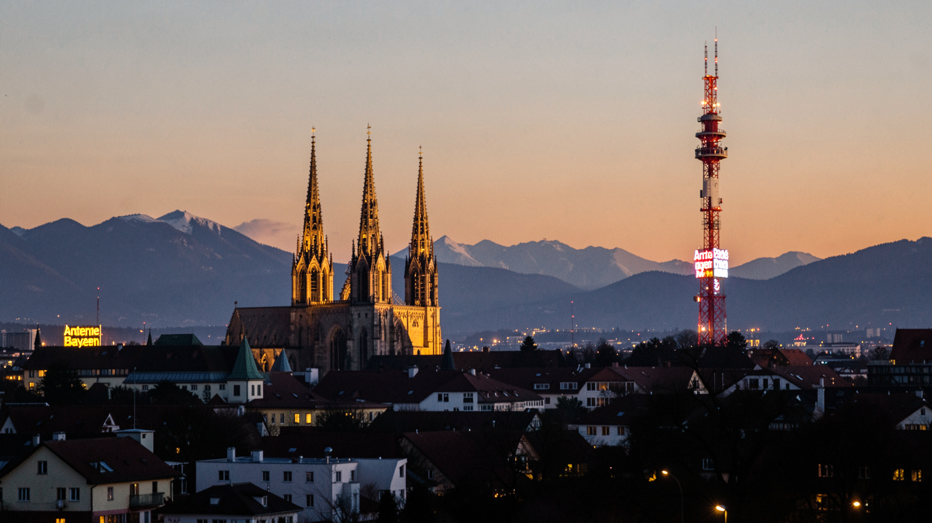 The Munich skyline at dusk with a broadcast tower sign reading Antenne Bayern and the Alps in the distance