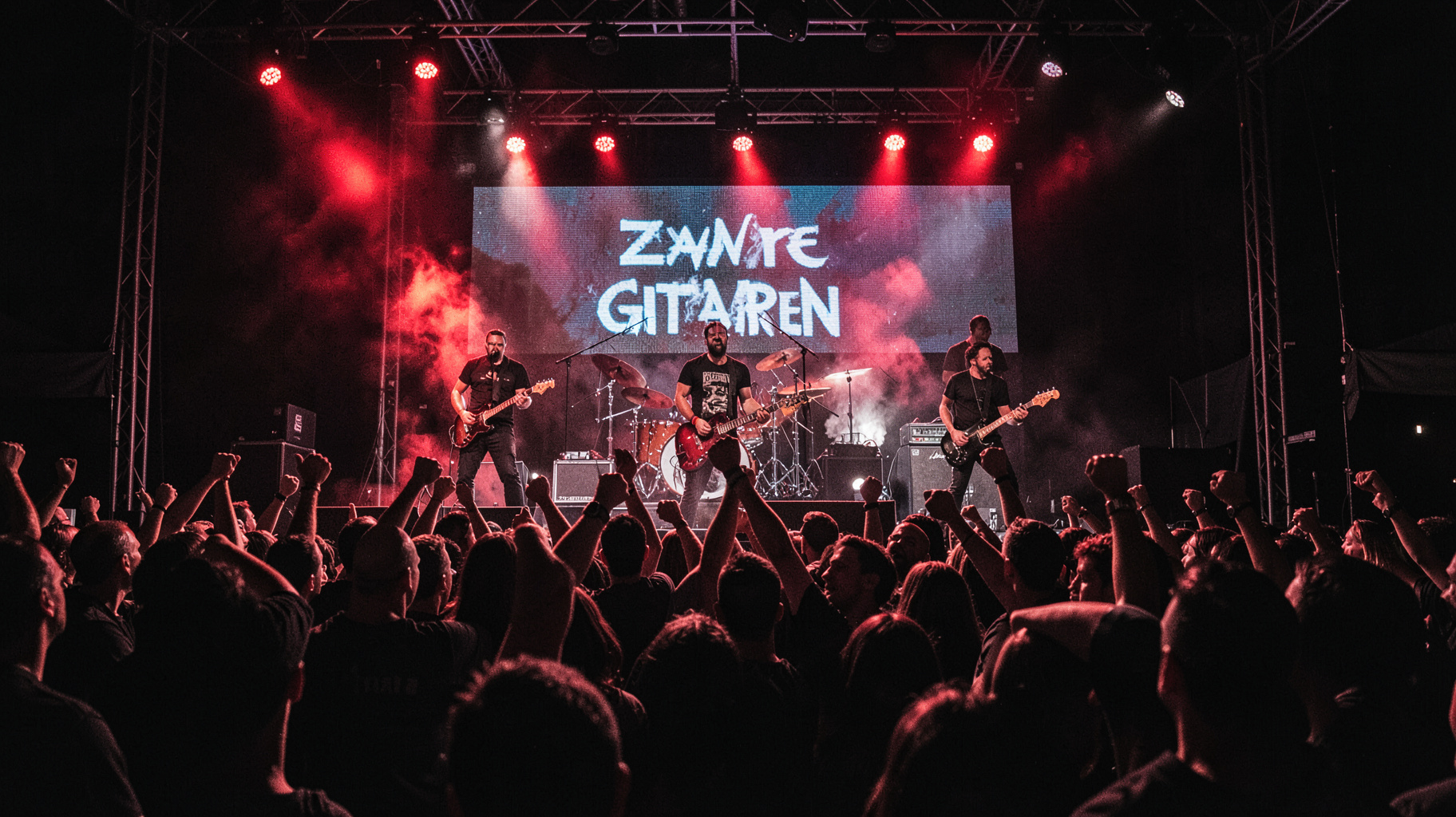 A dark heavy metal concert crowd with a screen behind the stage reading Zware Gitaren