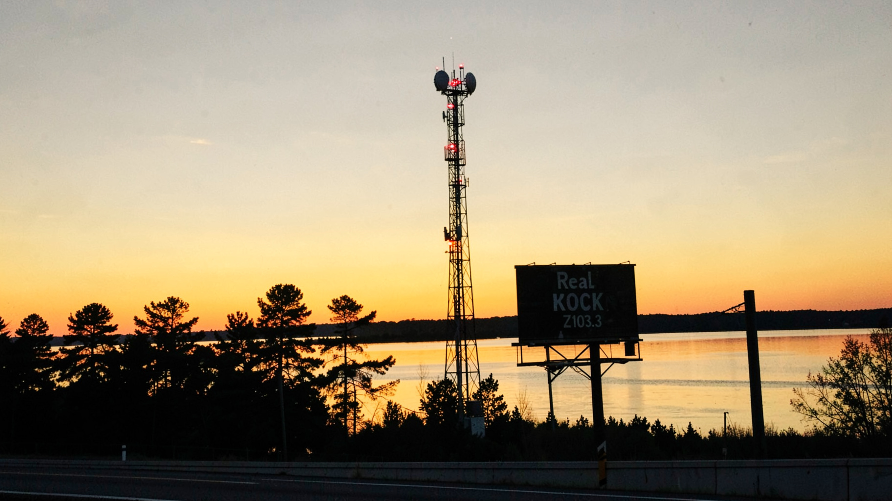 A radio broadcast tower silhouetted against a Minnesota lake sunset with a highway billboard reading Real Rock Z103.3