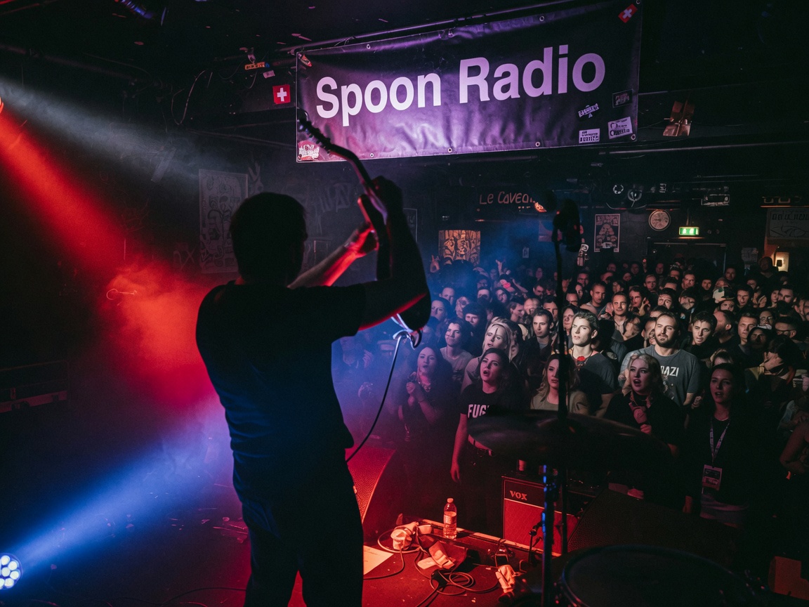 A moody indie rock concert crowd in a Geneva club with a Spoon Radio banner above the stage