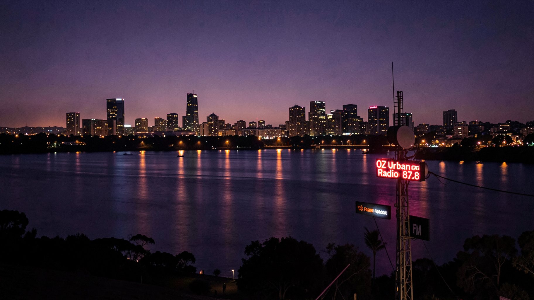 A nighttime Perth skyline along the Swan River with a glowing radio tower reading OZ Urban Radio 87.8
