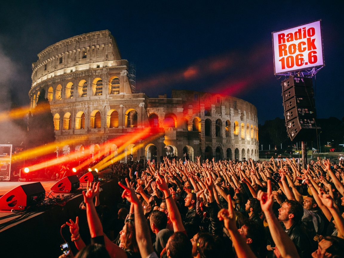 A rock concert crowd at night in Rome with the illuminated Colosseum in the background and a glowing Radio Rock 106.6 sign on the speaker stack