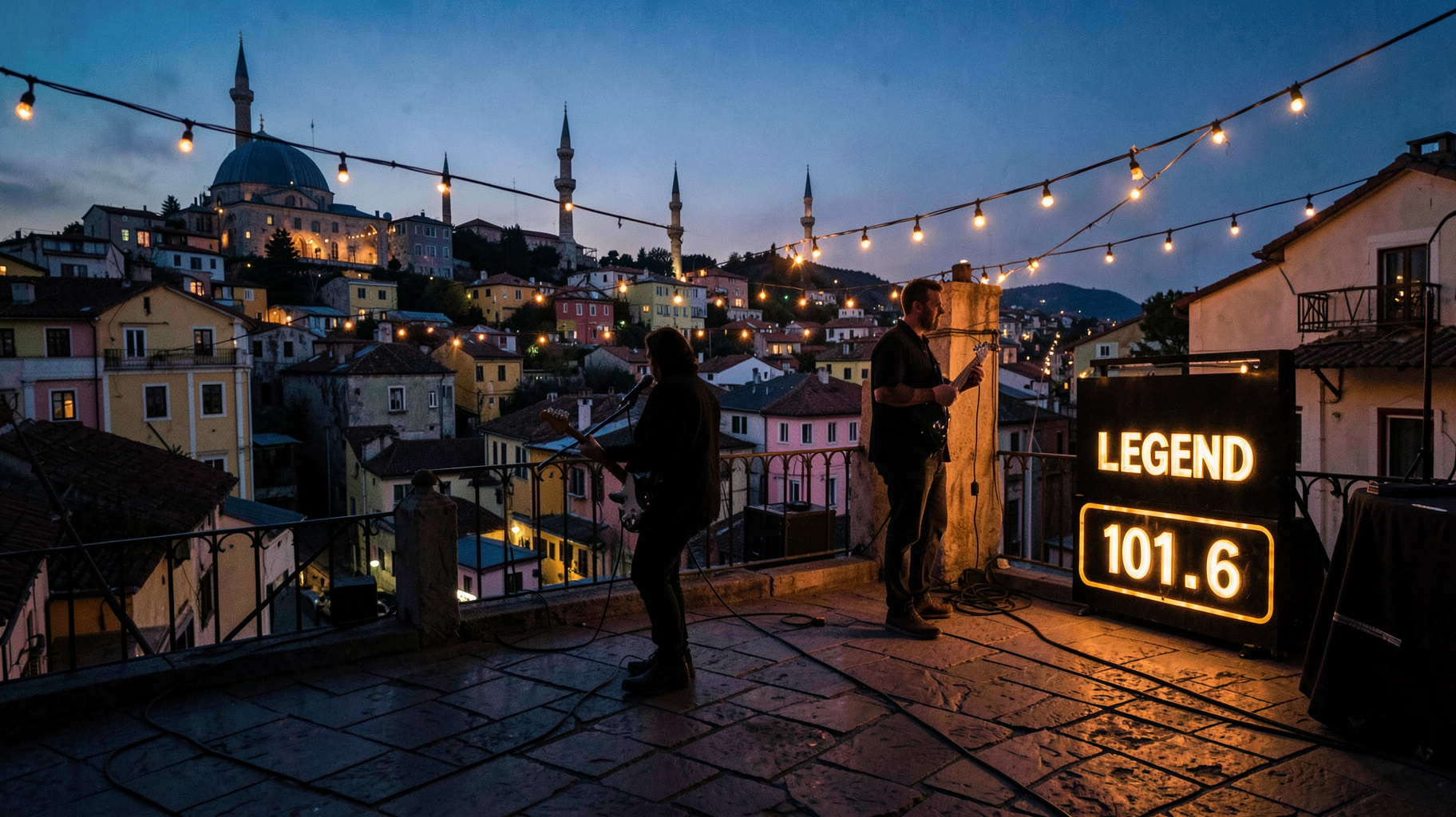 A rock guitarist silhouette on a rooftop stage above Xanthi's Old Town at dusk, with a marquee sign reading Legend 101.6