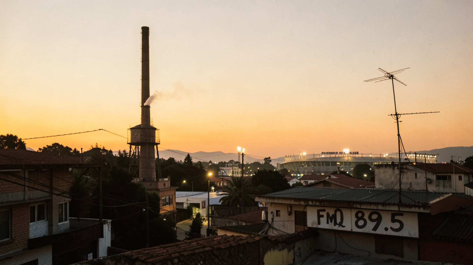 A golden hour streetscape in Quilmes, Argentina with a brewery smokestack, stadium lights, and a rooftop radio antenna marked FMQ