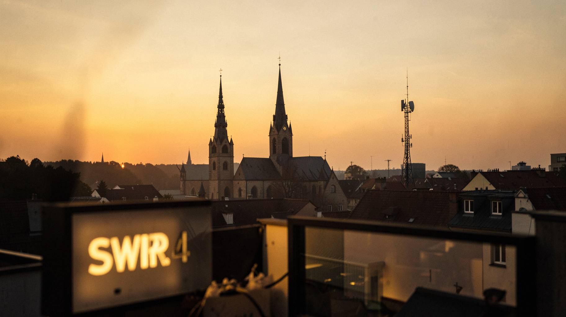 A warm golden-hour view over the rooftops of Kaiserslautern with a radio broadcast tower and a glowing SWR4 studio sign